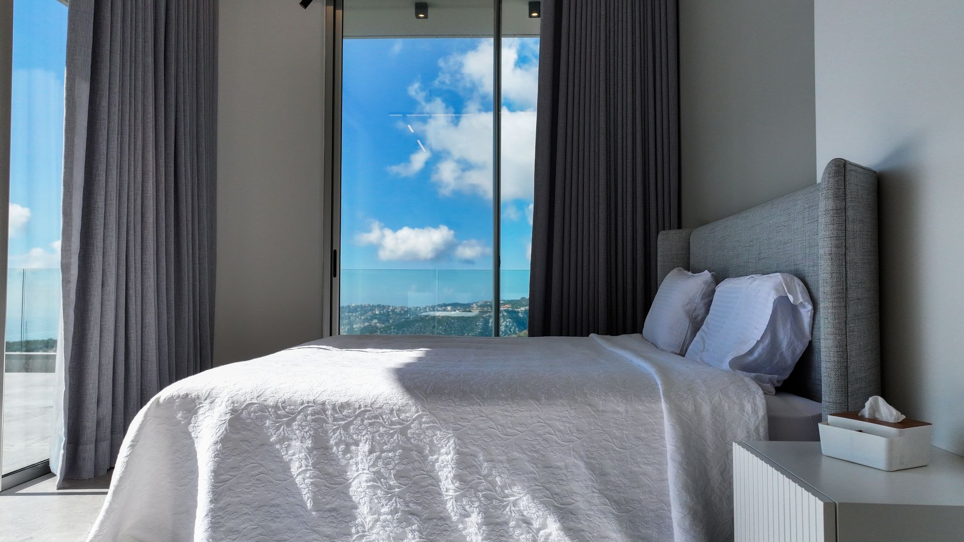 Bedroom with bed, window overlooking blue sky and ocean. Grey curtains, white bedding and nightstand.