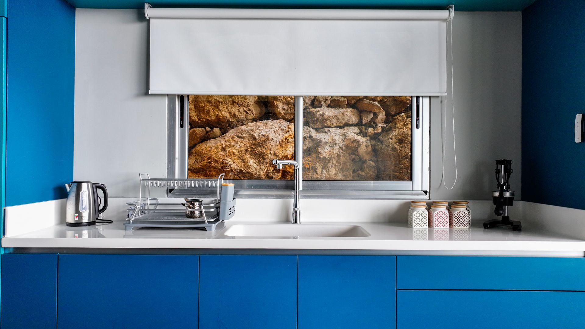 Blue kitchen with white countertop, window looking at rocks, silver kettle and dish rack.