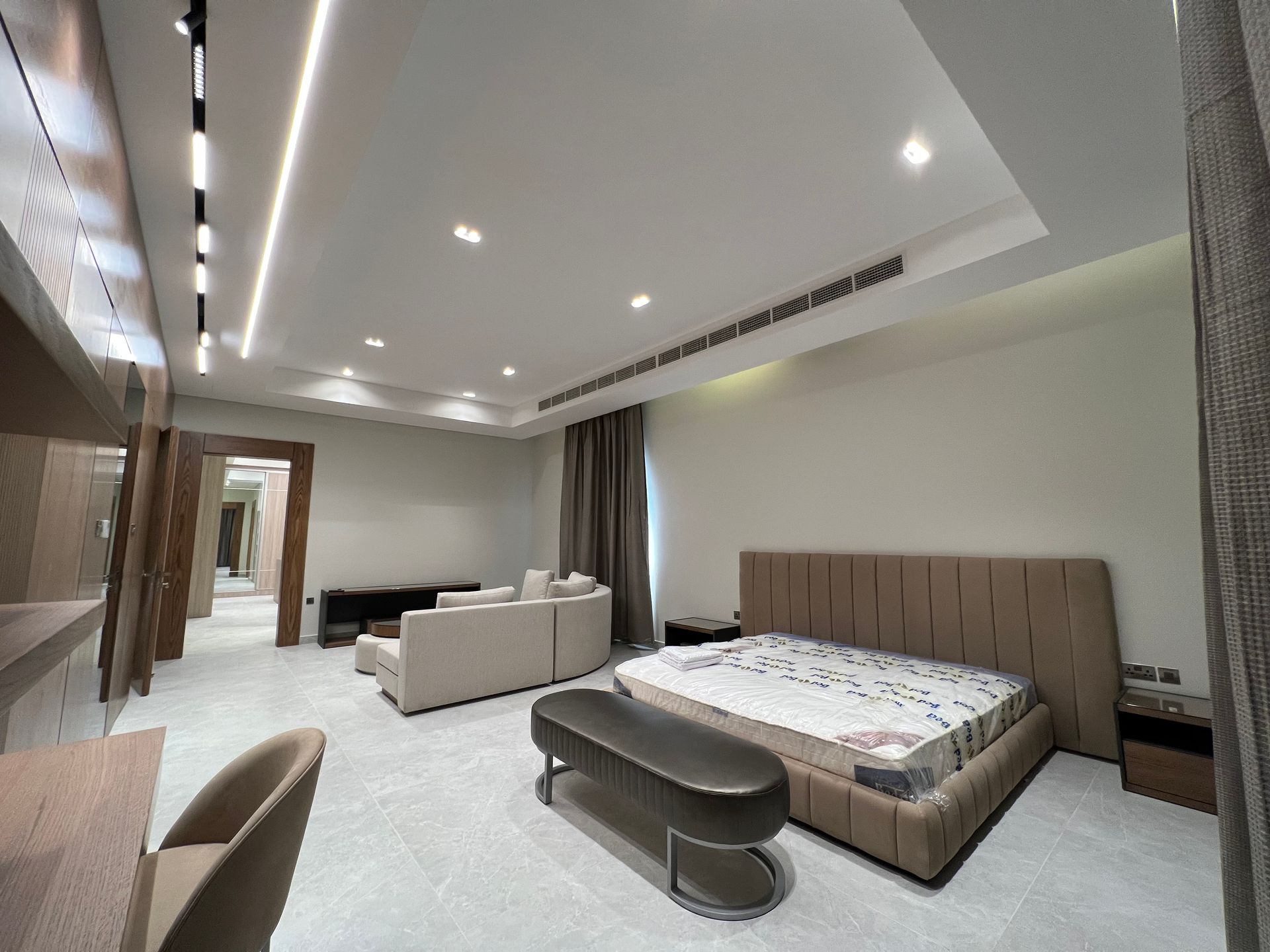 Spacious bedroom with a bed, sofa, desk, and modern lighting. Neutral colors dominate the room.