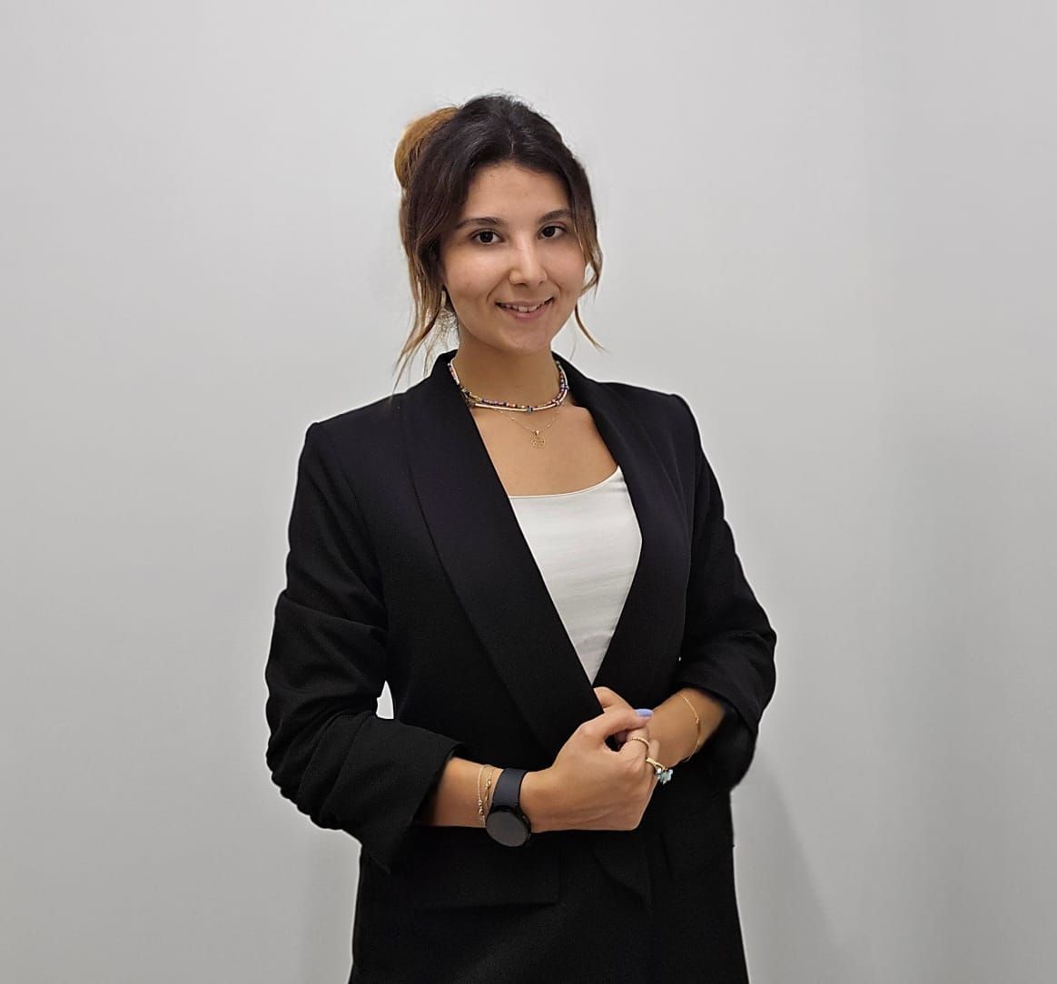 Woman in black blazer and white top smiles, standing against a white wall.