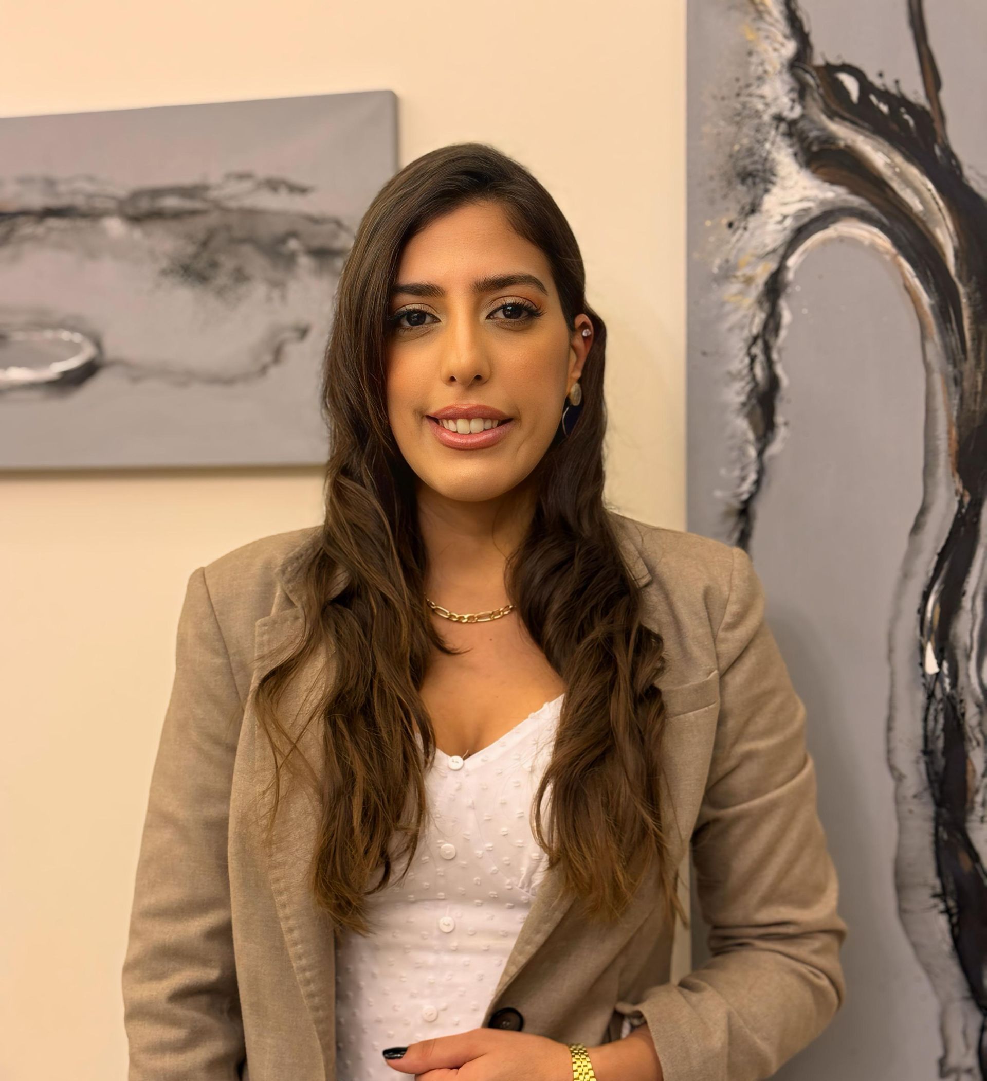 Woman with long brown hair, smiling, wearing a tan blazer and white top, in front of abstract art.