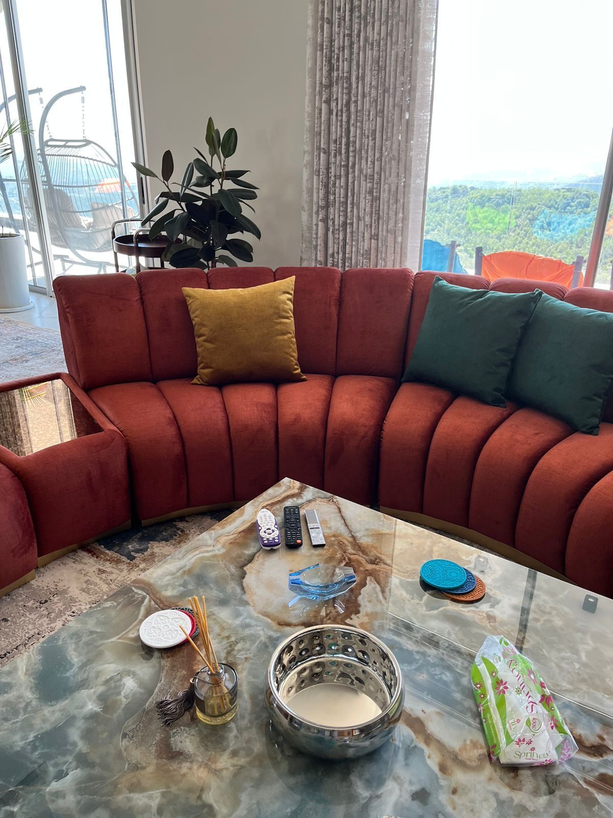 An orange velvet sectional sofa with gold trim and throw pillows sits next to a marble coffee table.