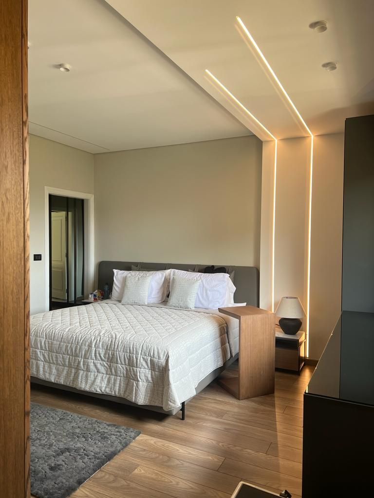 Bedroom with bed, neutral color walls, wooden floor, and linear ceiling lights.