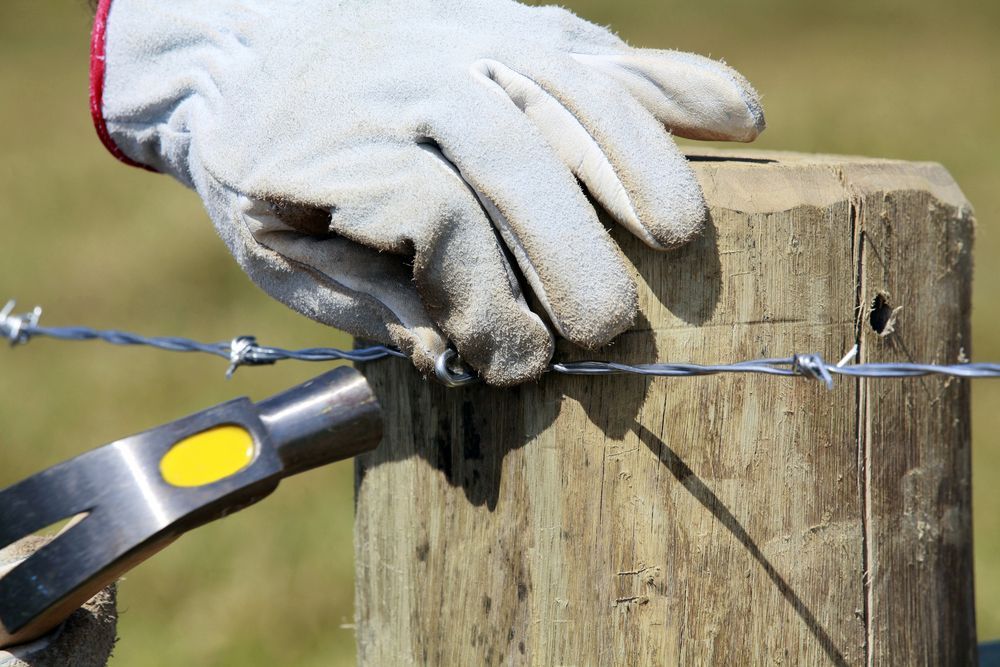 A Person is Using a Hammer to Nail Barbed Wire to a Wooden Post — Perry's Timber & Fencing In Shellharbour, NSW