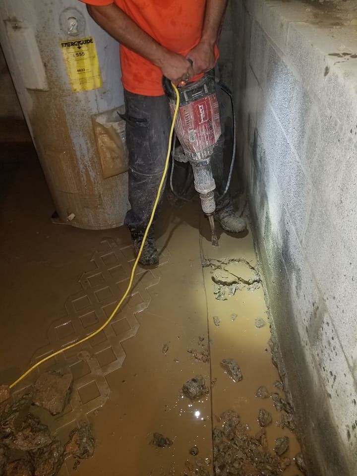 A person in an orange shirt using a jackhammer to break up concrete in a flooded basement.