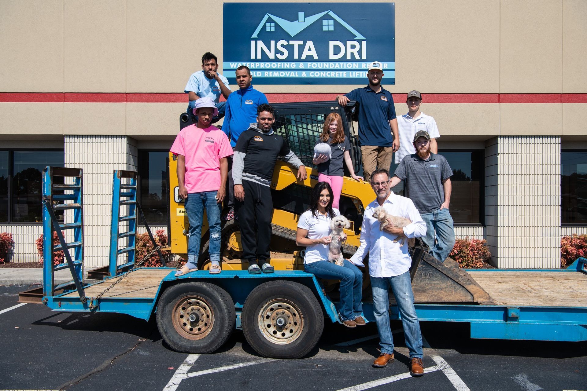 Insta-Dri team posing on a trailer with a small excavator, outdoors. They are smiling in front of a building.