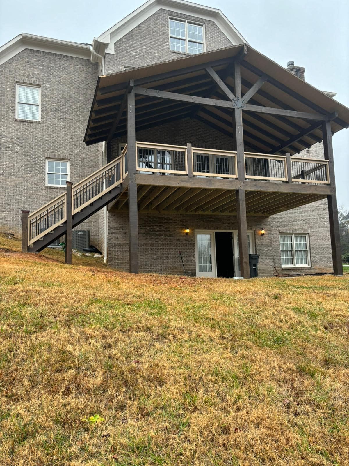 Back of a brick house with a wooden deck and covered patio, brown and gray tones on a grassy hill.
