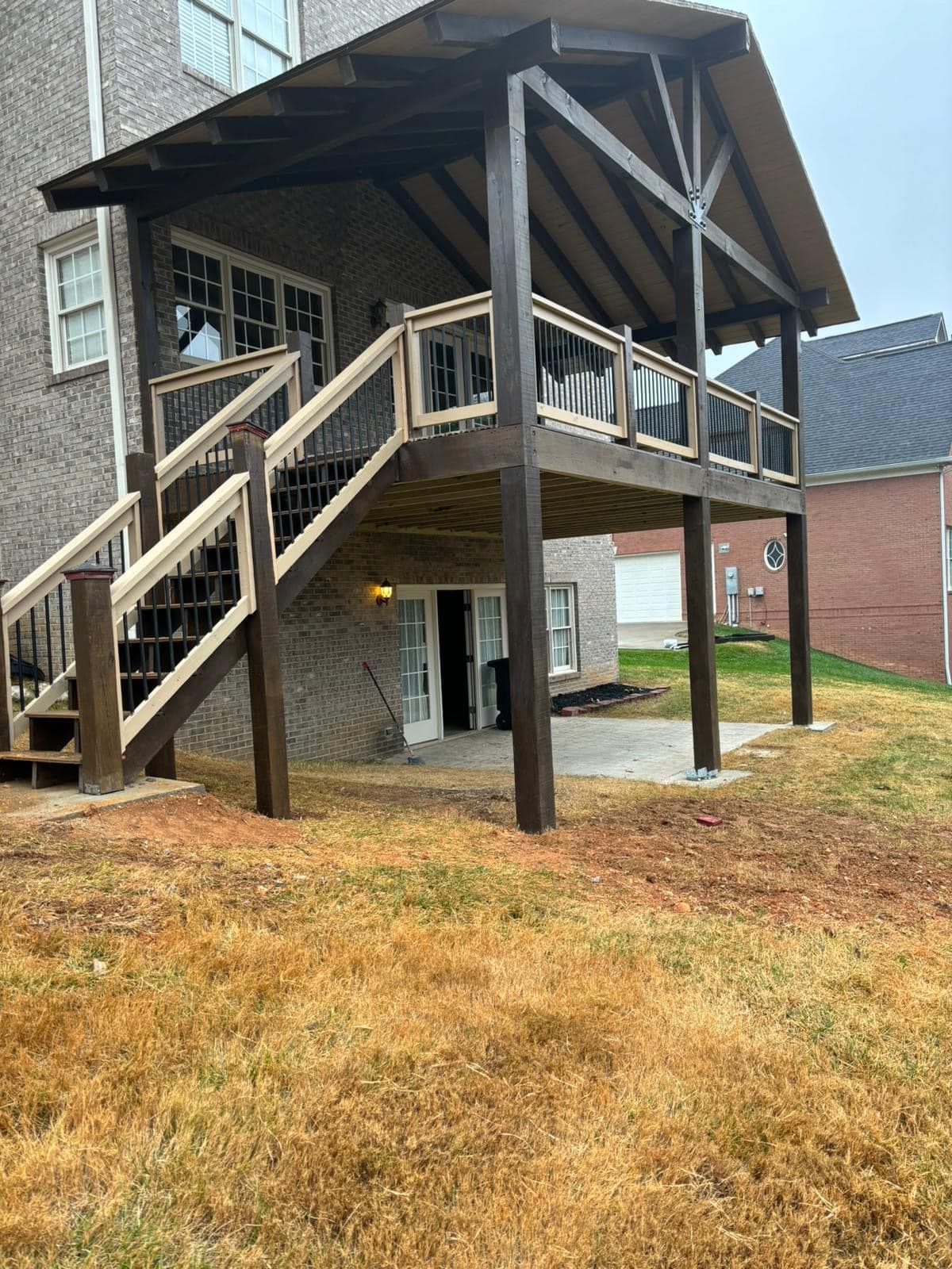 Wooden deck with stairs and a covered porch, brown and beige, next to a brick house and grass.