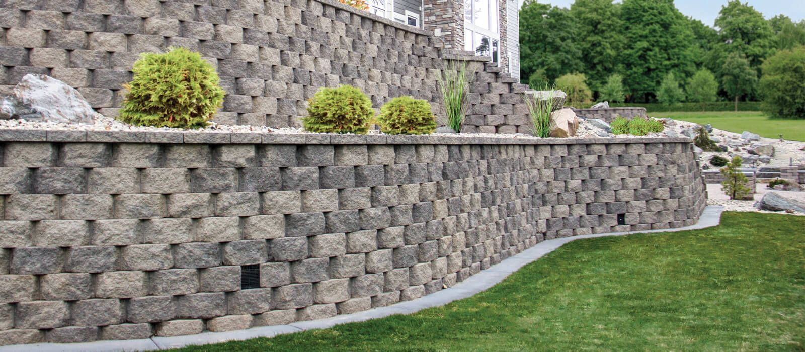 Stone retaining wall with planted bushes, beside a green lawn.