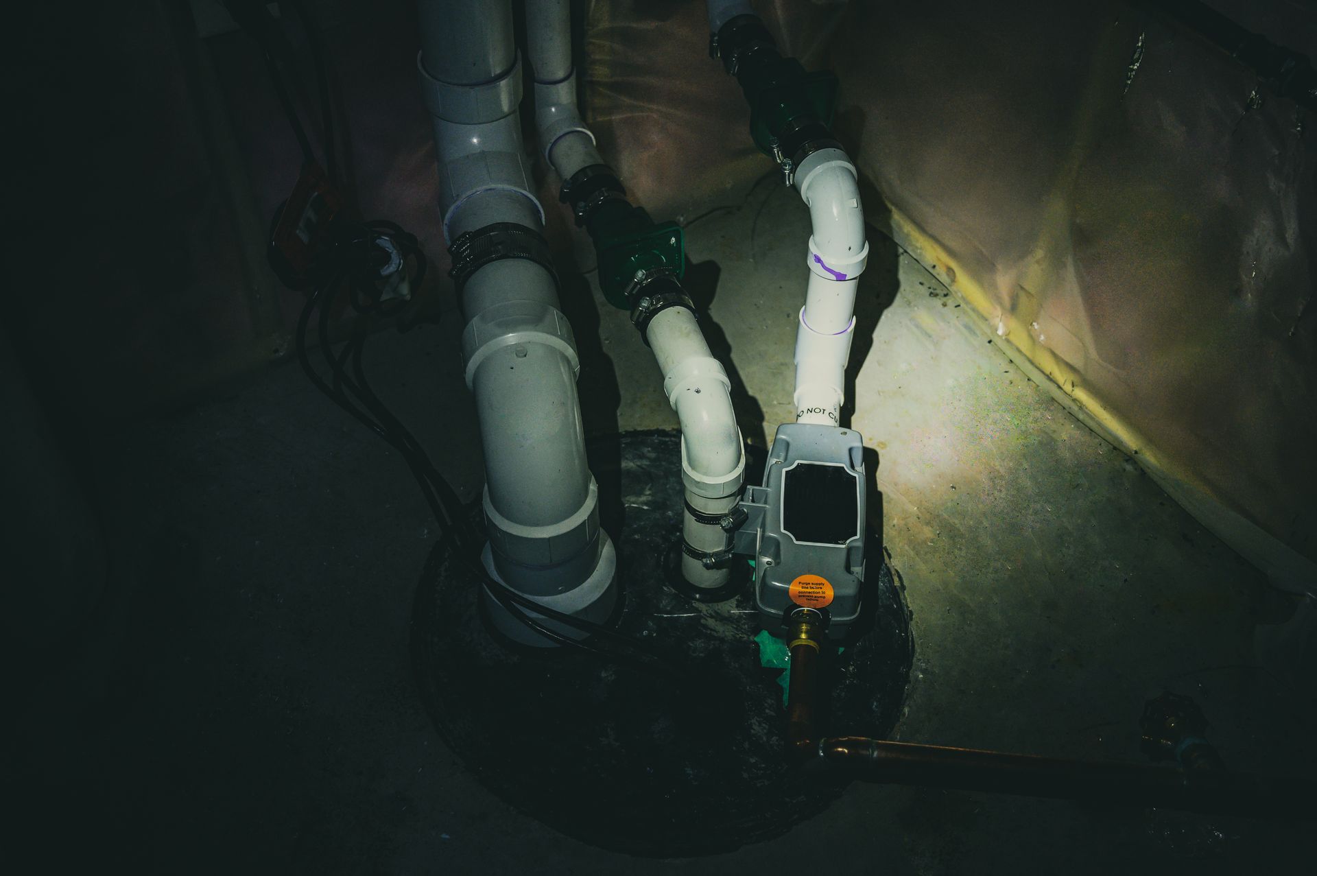 Sump pump system with gray and white pipes in a dark basement corner.