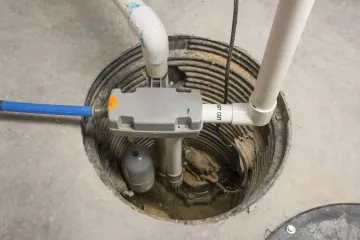 Open utility access hole with pipes, wiring, and a gray device mounted over the opening.
