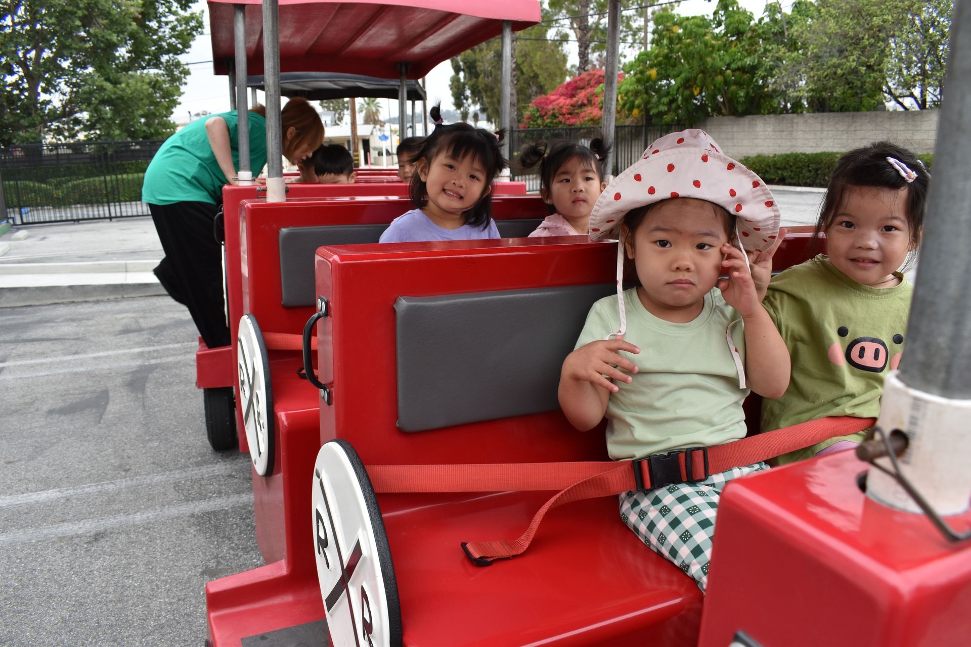 Children riding a red toy train, smiling. Outdoors, person in green shirt operating train.