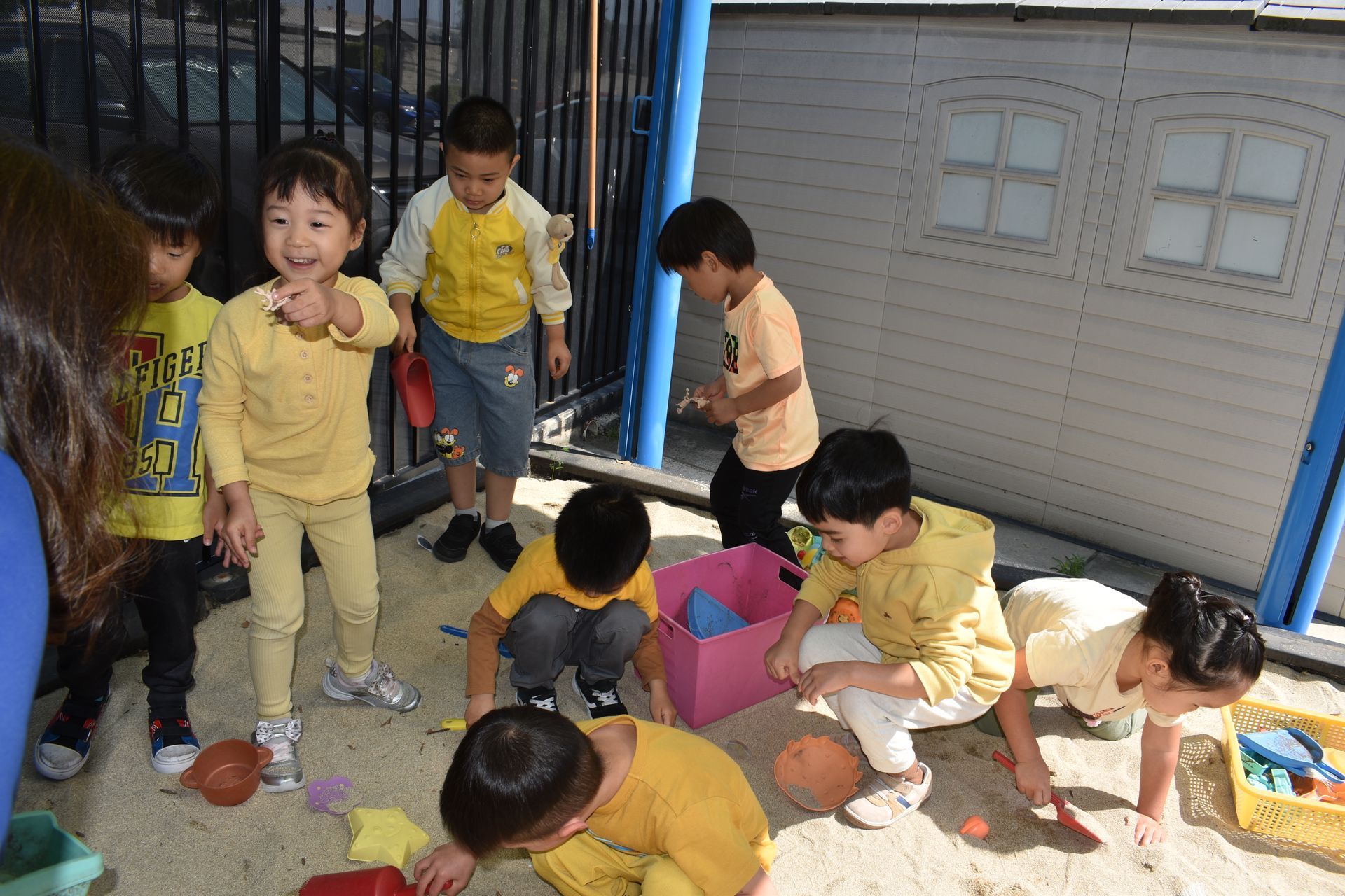 Children playing in sandbox, some wearing yellow, near a playhouse.