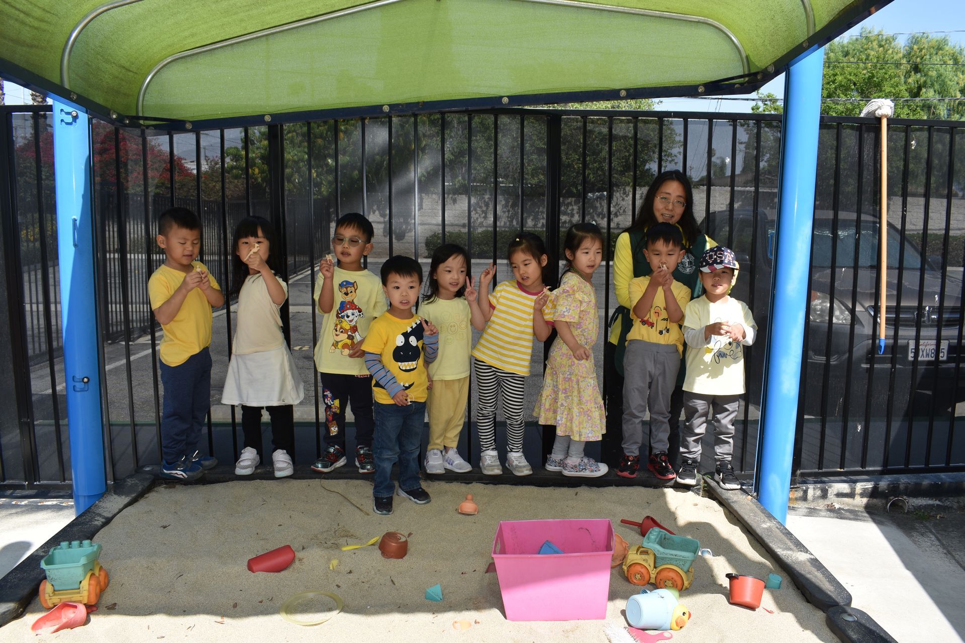 Children and teacher in yellow clothing posing under shade structure in sandbox.