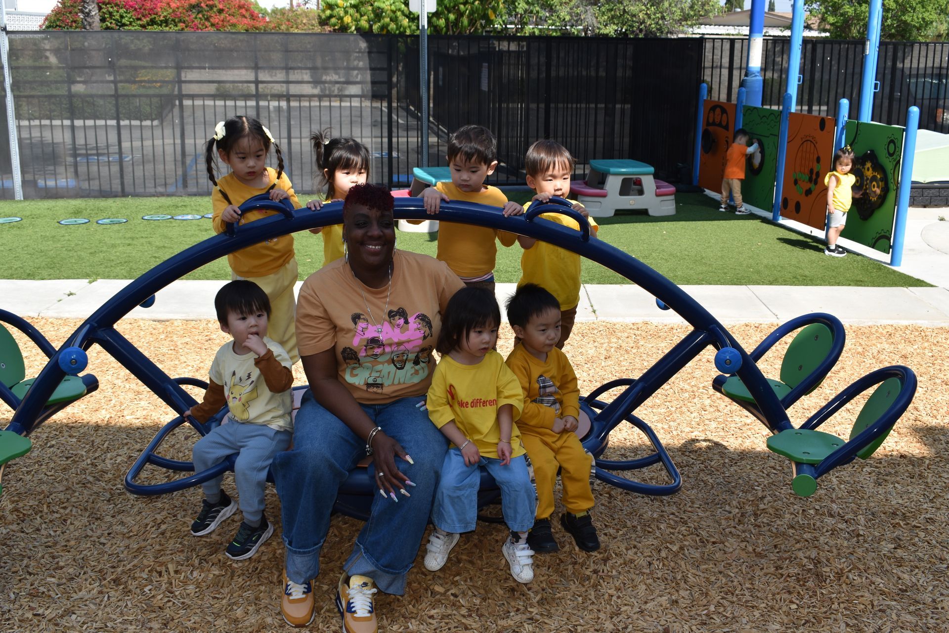 A teacher and children in yellow shirts on a playground seesaw, smiles.