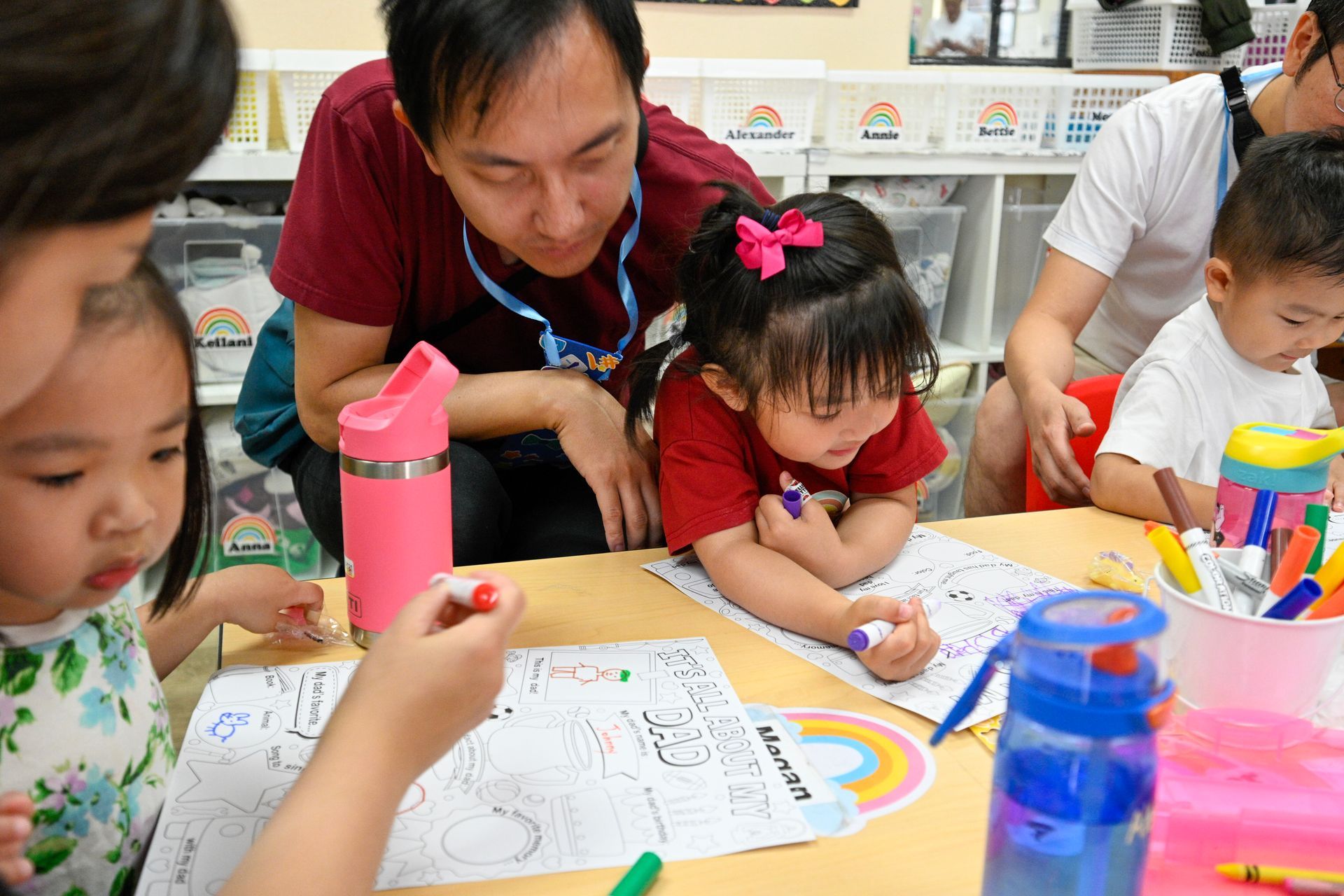 Adults helping young children color at a table in a brightly lit classroom.
