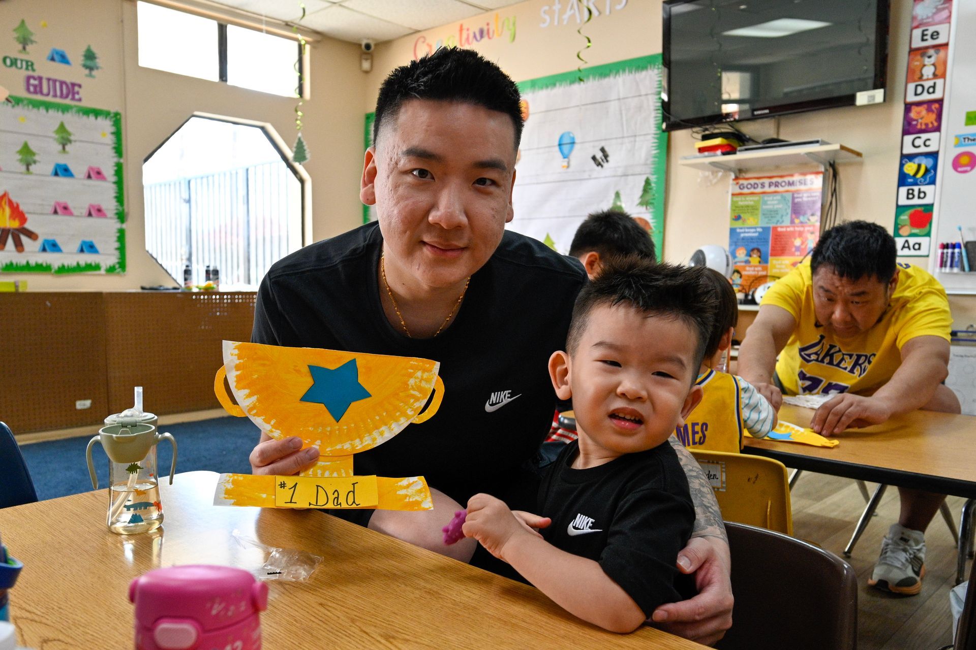 Man and child holding a paper plate trophy in a classroom. Other children crafting at tables.