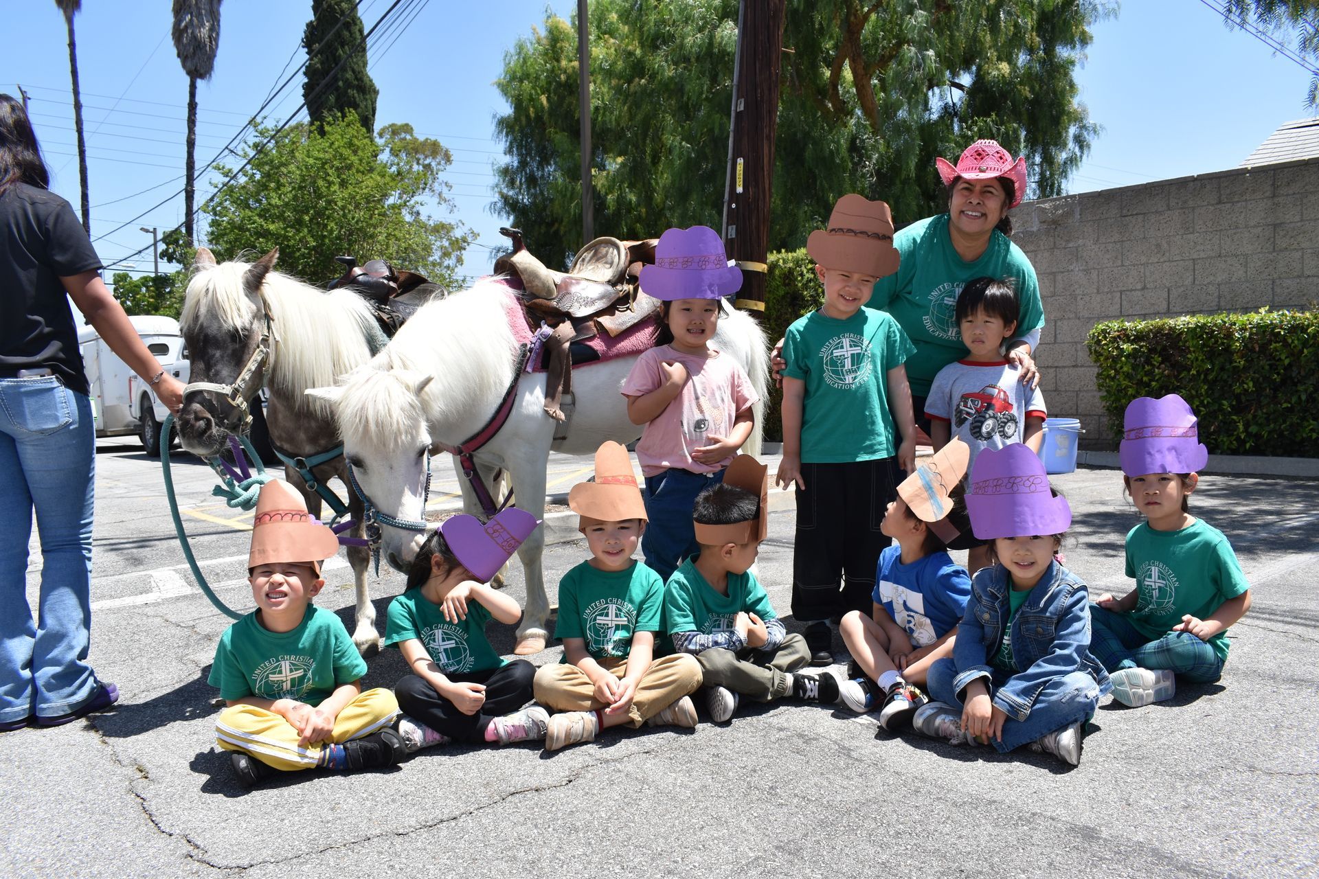 Group of kids in green shirts and paper hats pose with ponies outdoors.