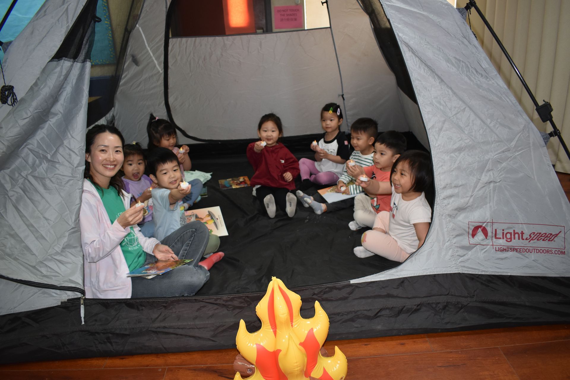Children and a teacher in a tent, pretending to camp. A stuffed campfire is in front.