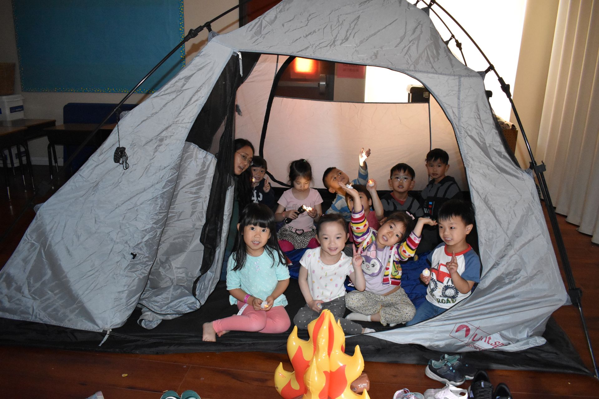 Children in a camping tent, smiling and holding up objects, with a toy campfire in front.