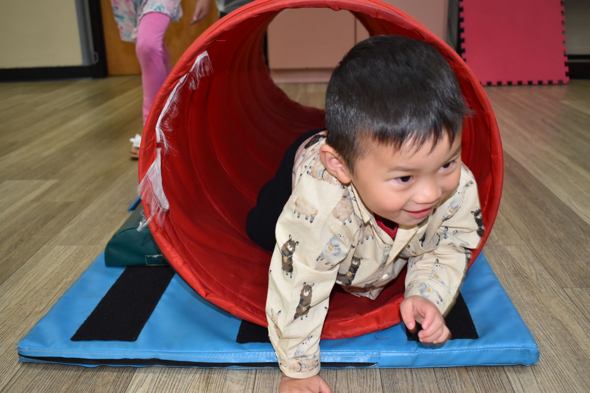 Young child crawling through a red tunnel on a blue mat, smiling.