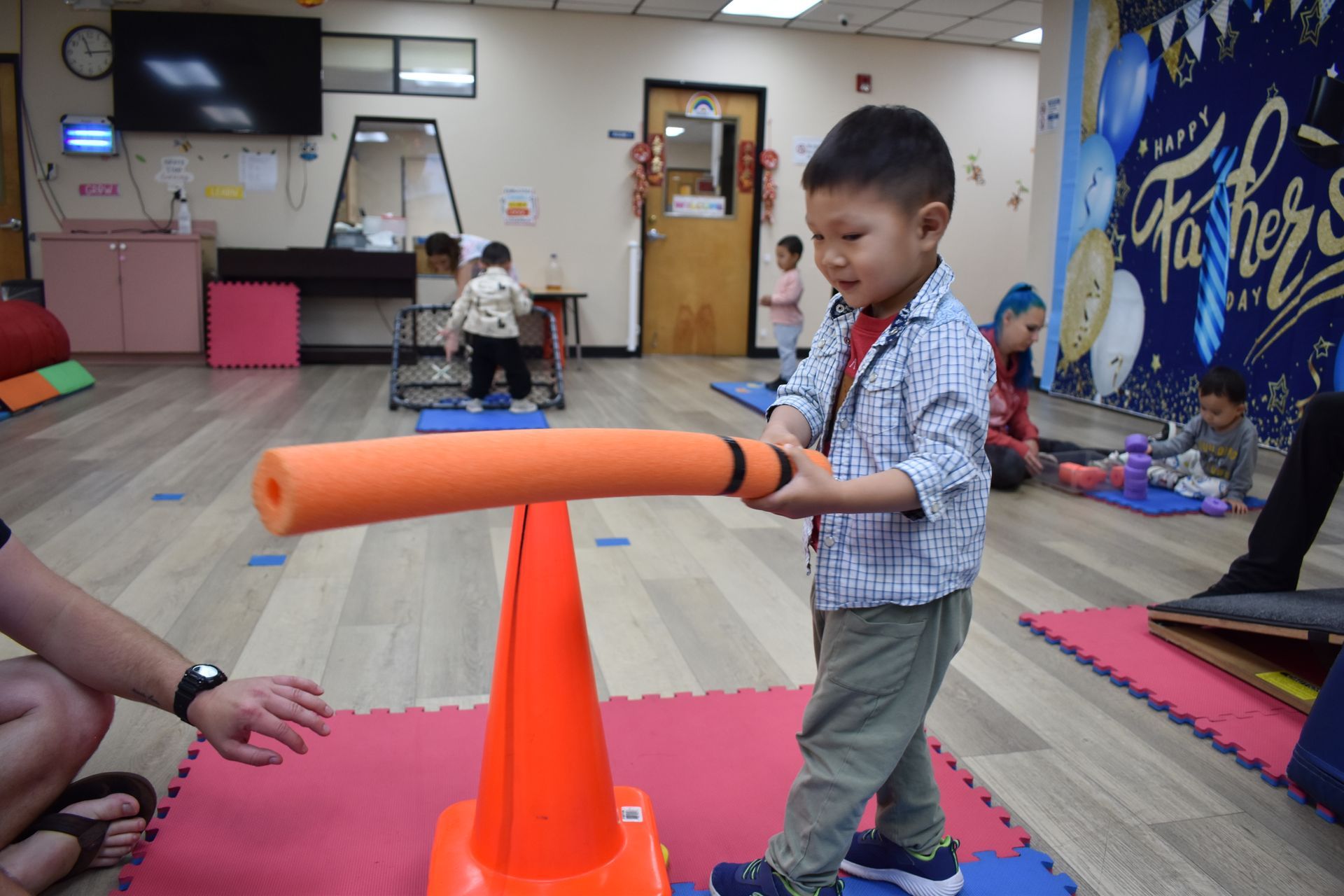 Boy balances pool noodle on a traffic cone, smiles. Indoors, other children play.