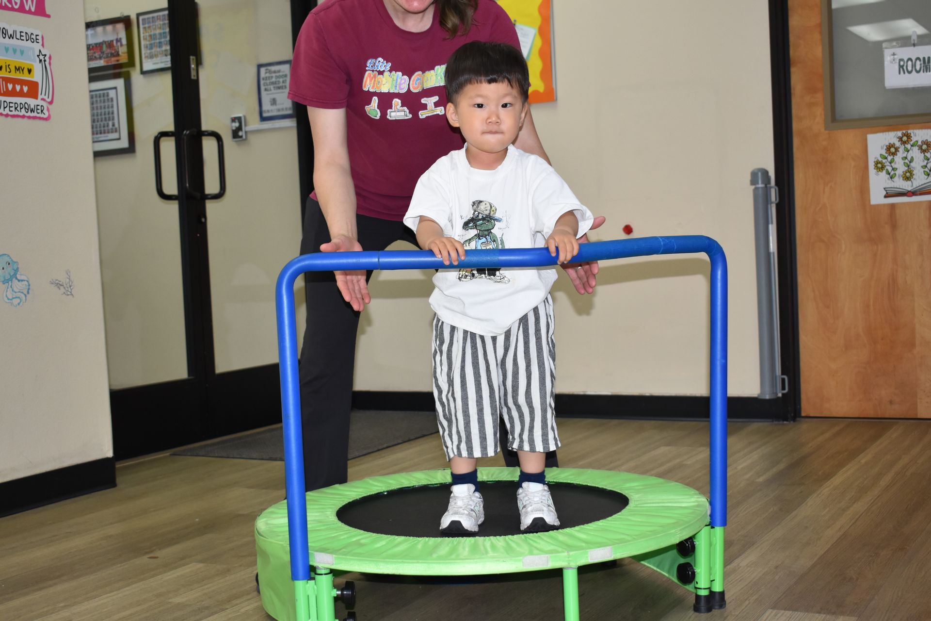 Child on a mini trampoline with a handle, being supported by an adult indoors.