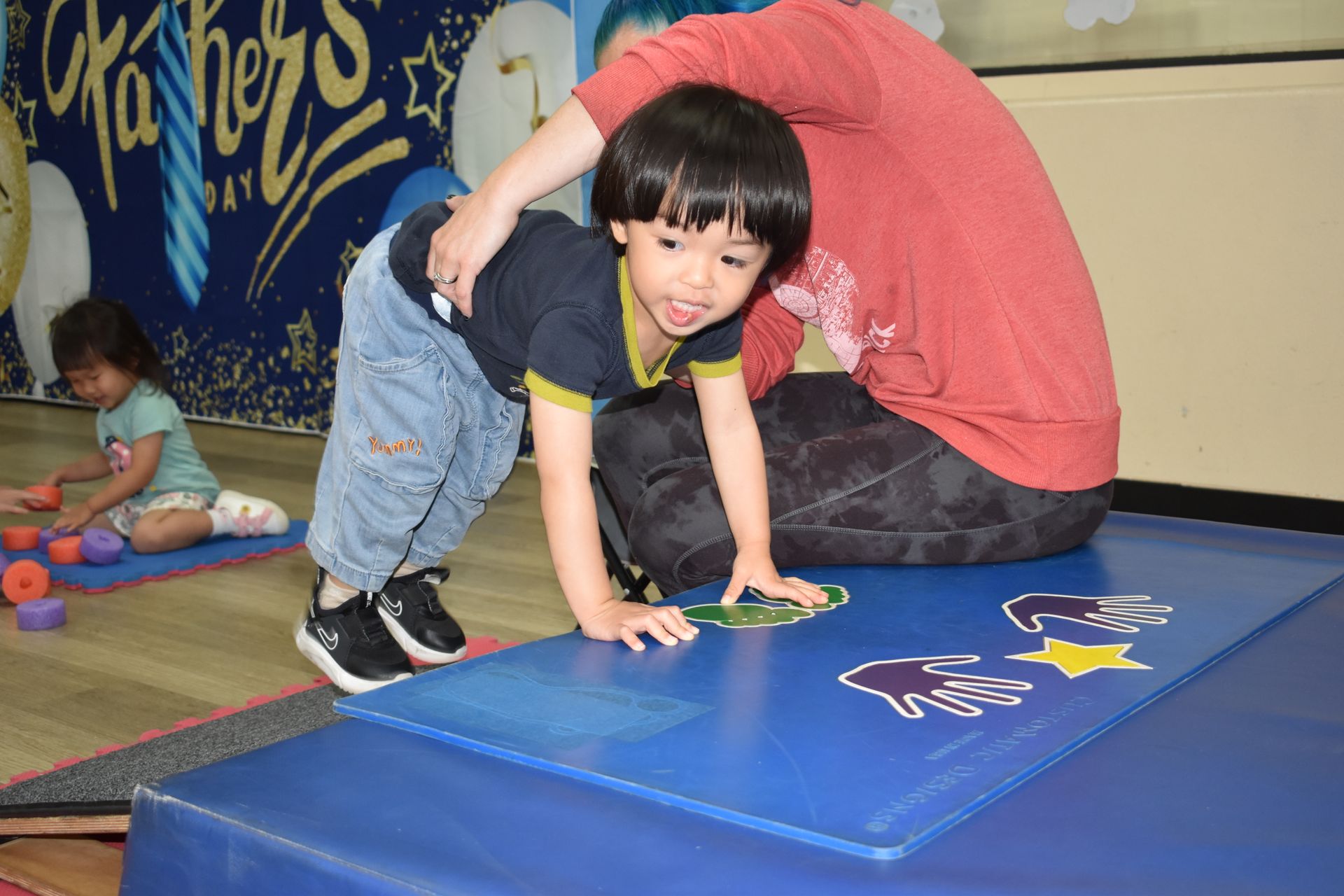 A young boy doing an exercise with assistance from an adult in a child's playroom.