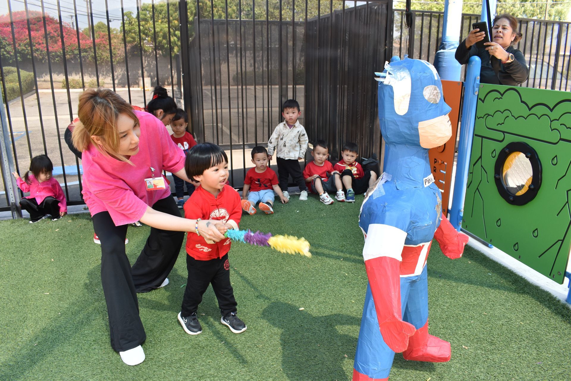 Woman and child hitting a Captain America piñata with kids watching outdoors.