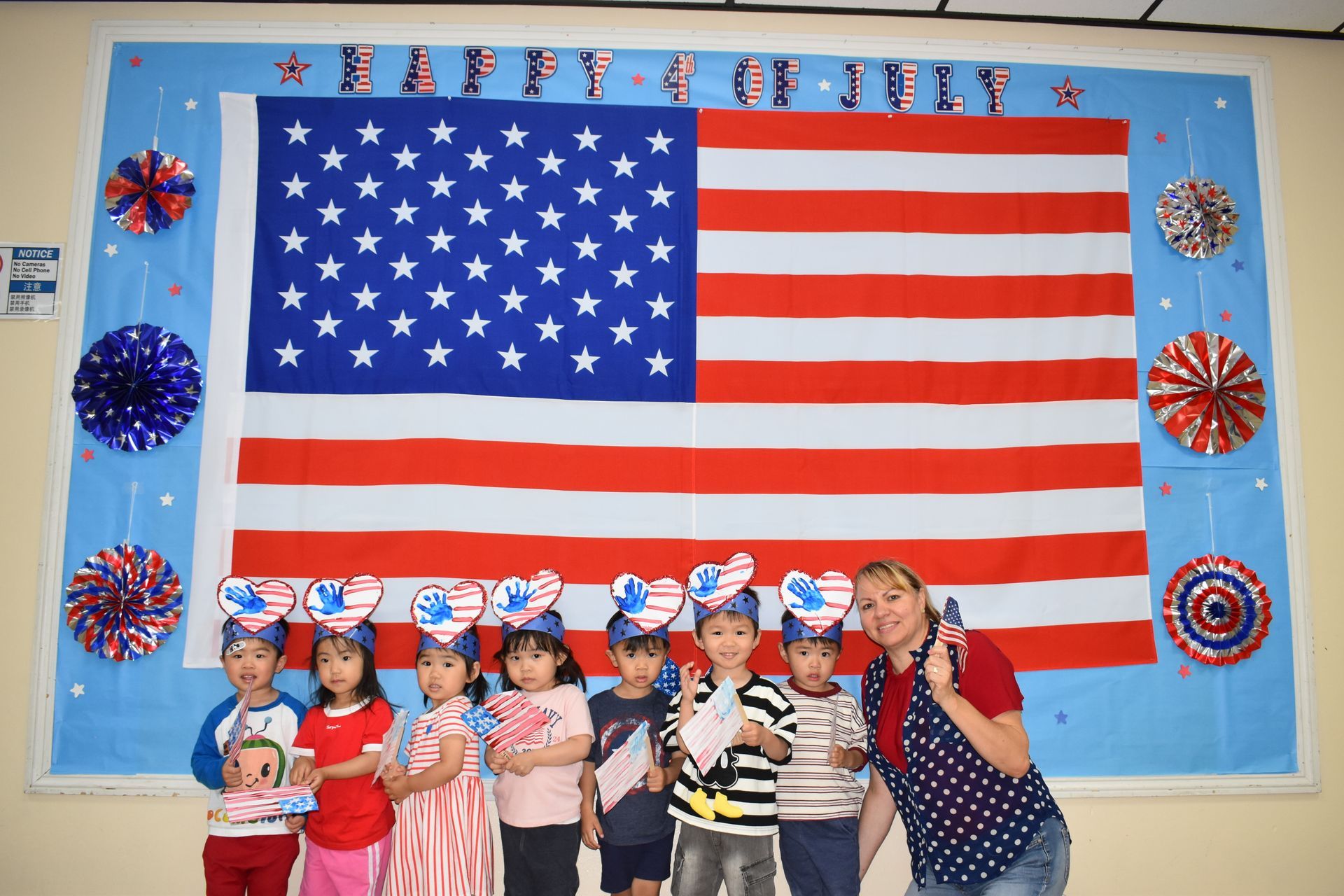 Children and teacher pose in front of a US flag, wearing patriotic headbands for a July 4th celebration.