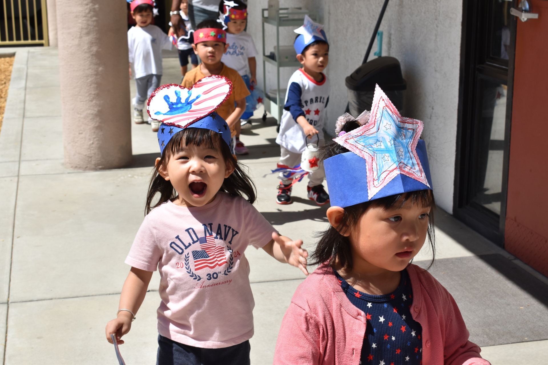 Children wearing patriotic hats walk outside, smiling and laughing on a sunny day.