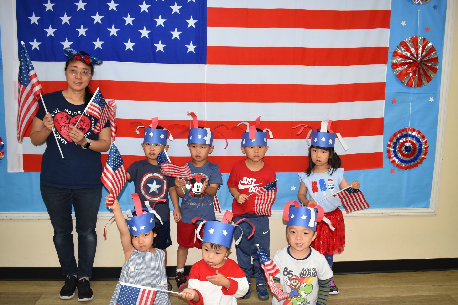Teacher and children posing in front of an American flag backdrop, holding flags, wearing patriotic hats.