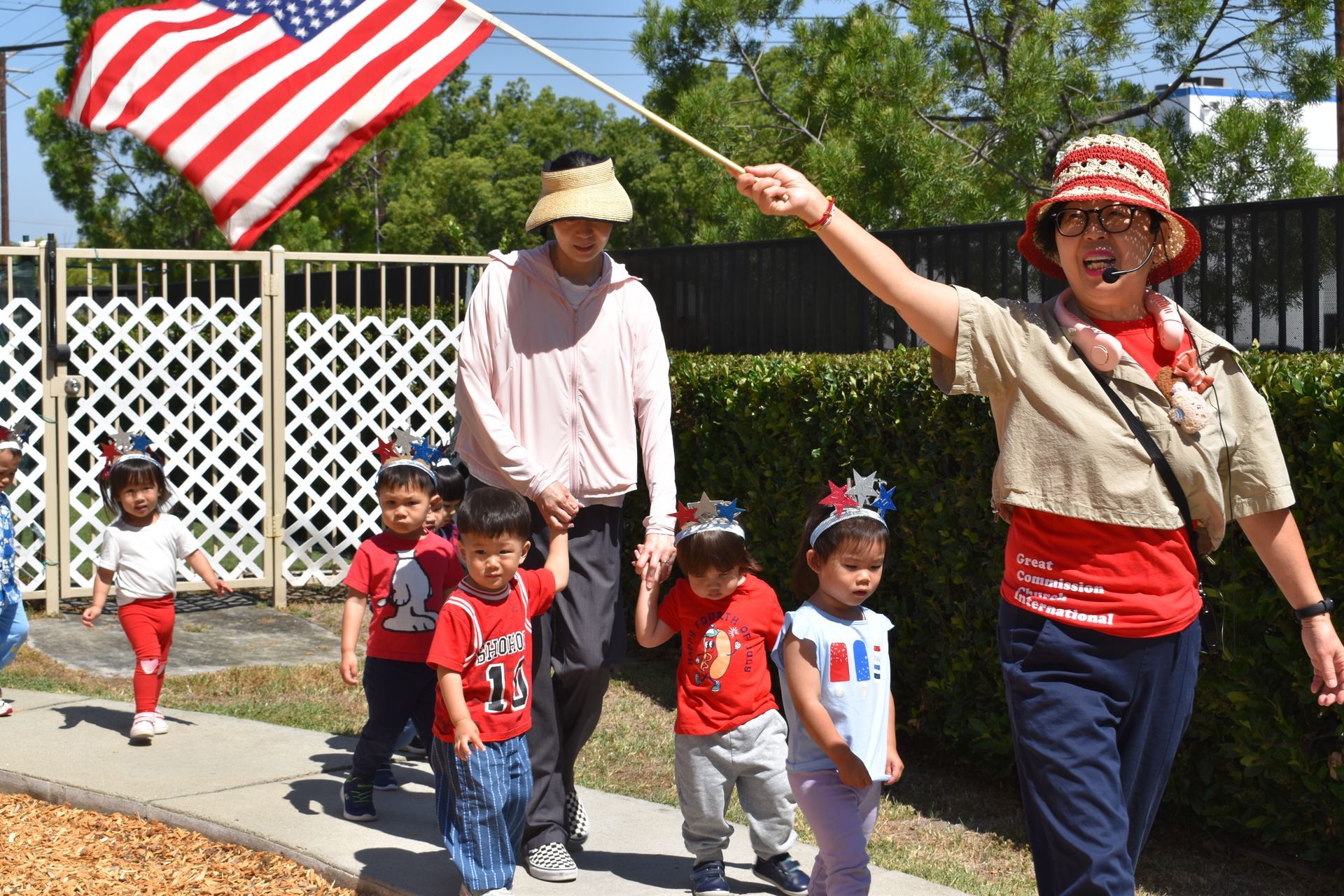 Children in red, white, and blue parade with adults carrying an American flag. Outdoors on a sunny day.