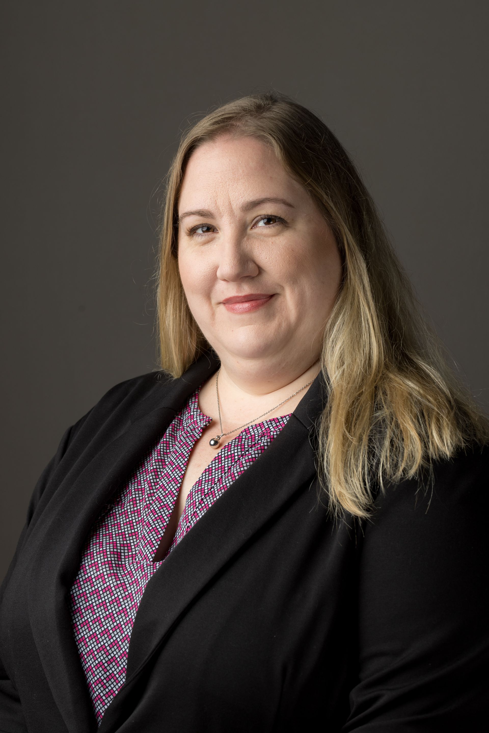 Woman with long blonde hair, wearing a blazer and patterned shirt, smiles at the camera.