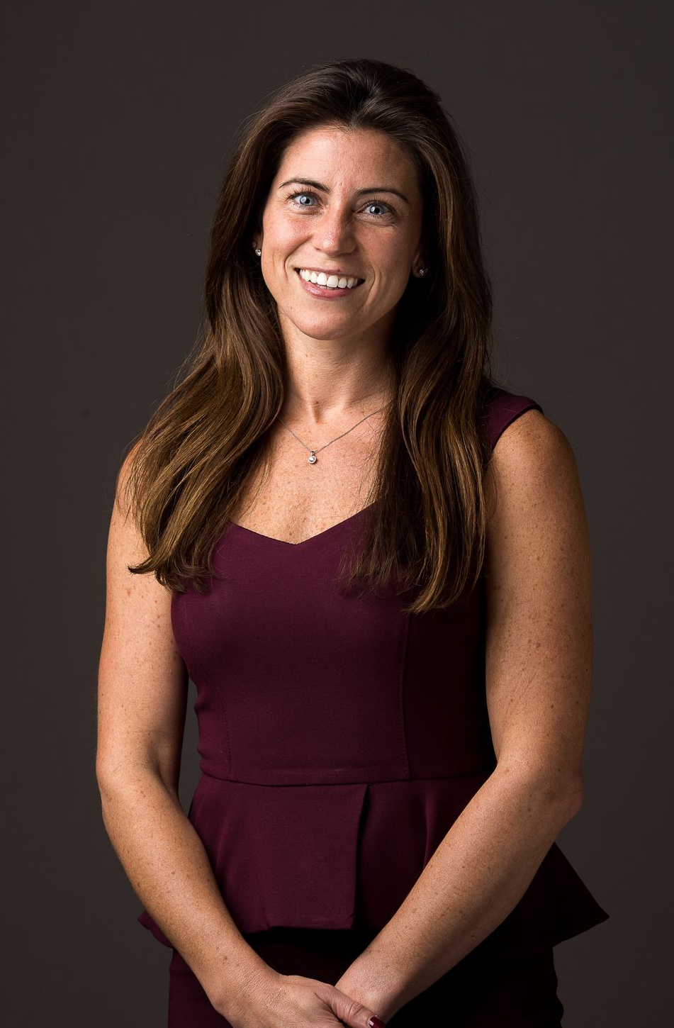Woman with brown hair smiles, wearing a sleeveless maroon top against a dark grey background.