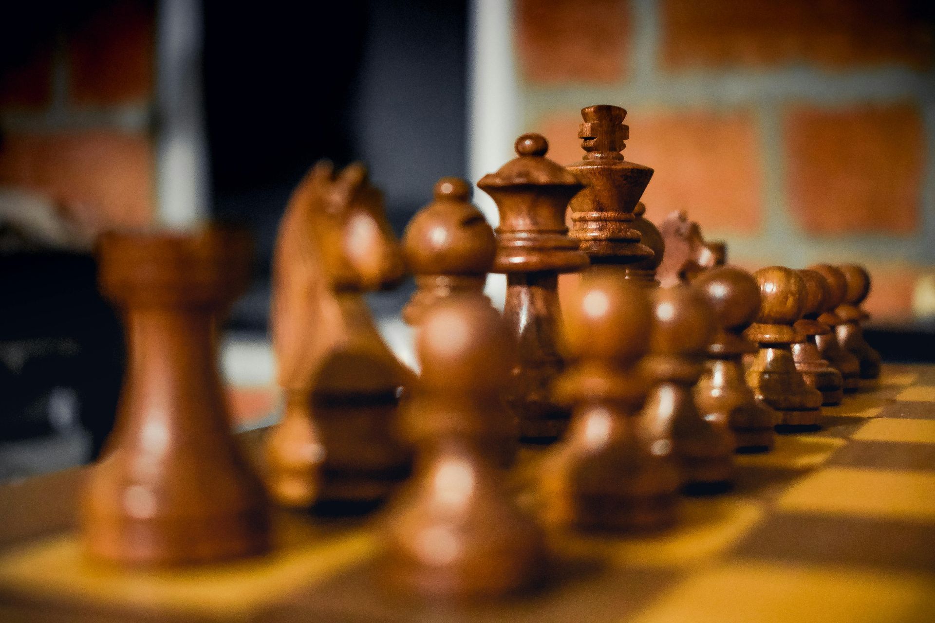 Chess pieces lined up on a board, focused on the back row. Brown pieces against a brick wall.