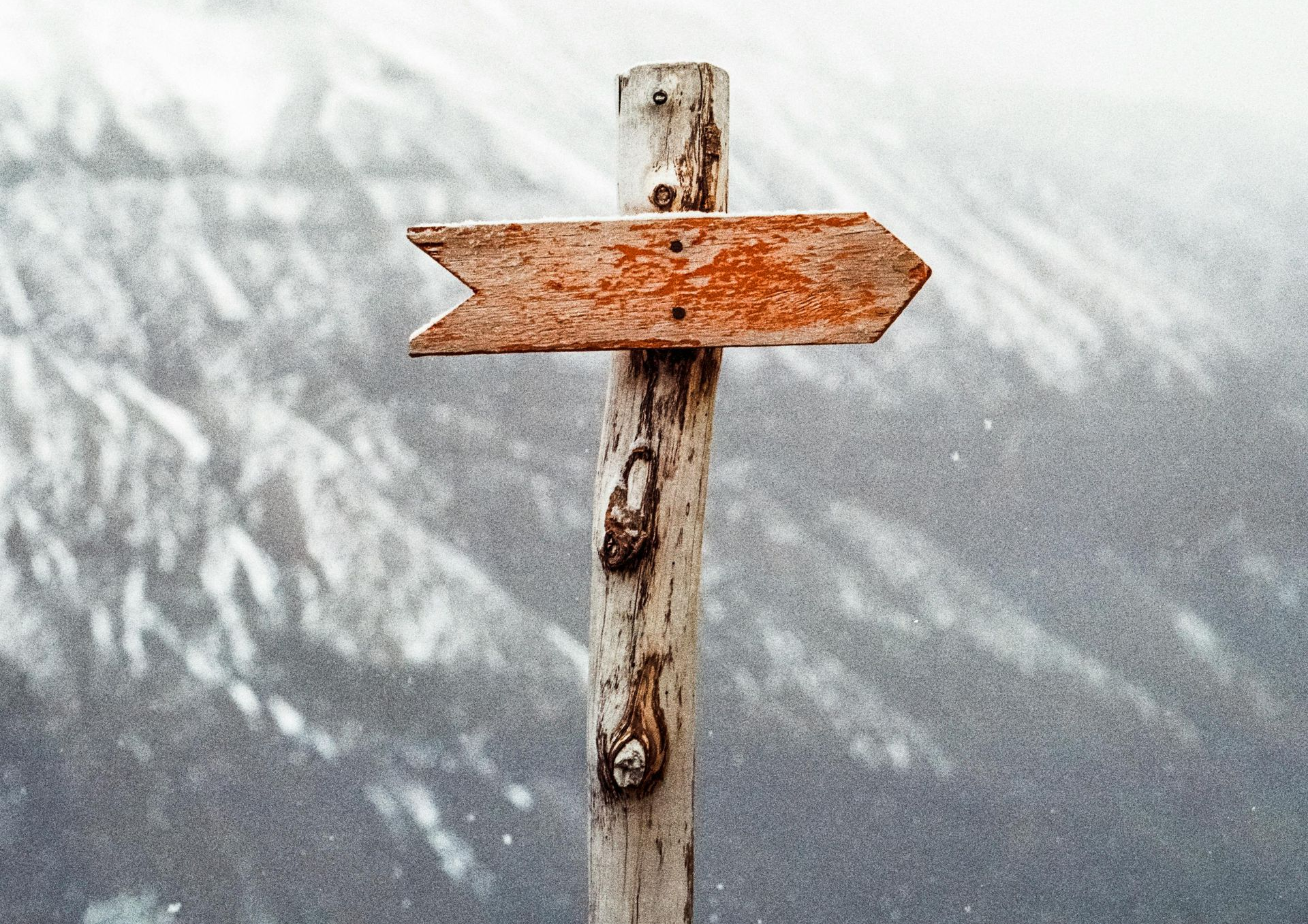Wooden signpost pointing right, orange arrow on weathered post, snowy mountain backdrop.