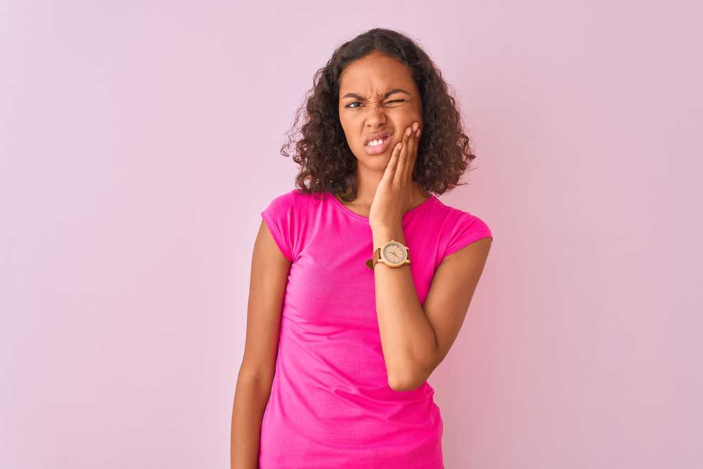 A young woman in a pink shirt is holding her face in pain because of a toothache.