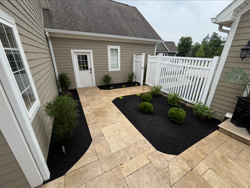 Tan stone patio with black mulch flowerbeds, shrubs, and white fence.