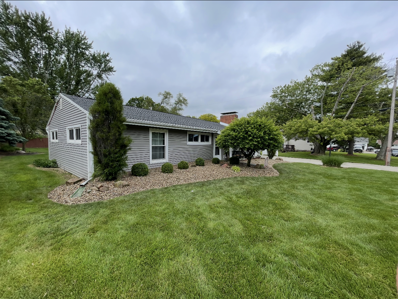 Gray house with white trim, green lawn, and rock landscaping under a cloudy sky.