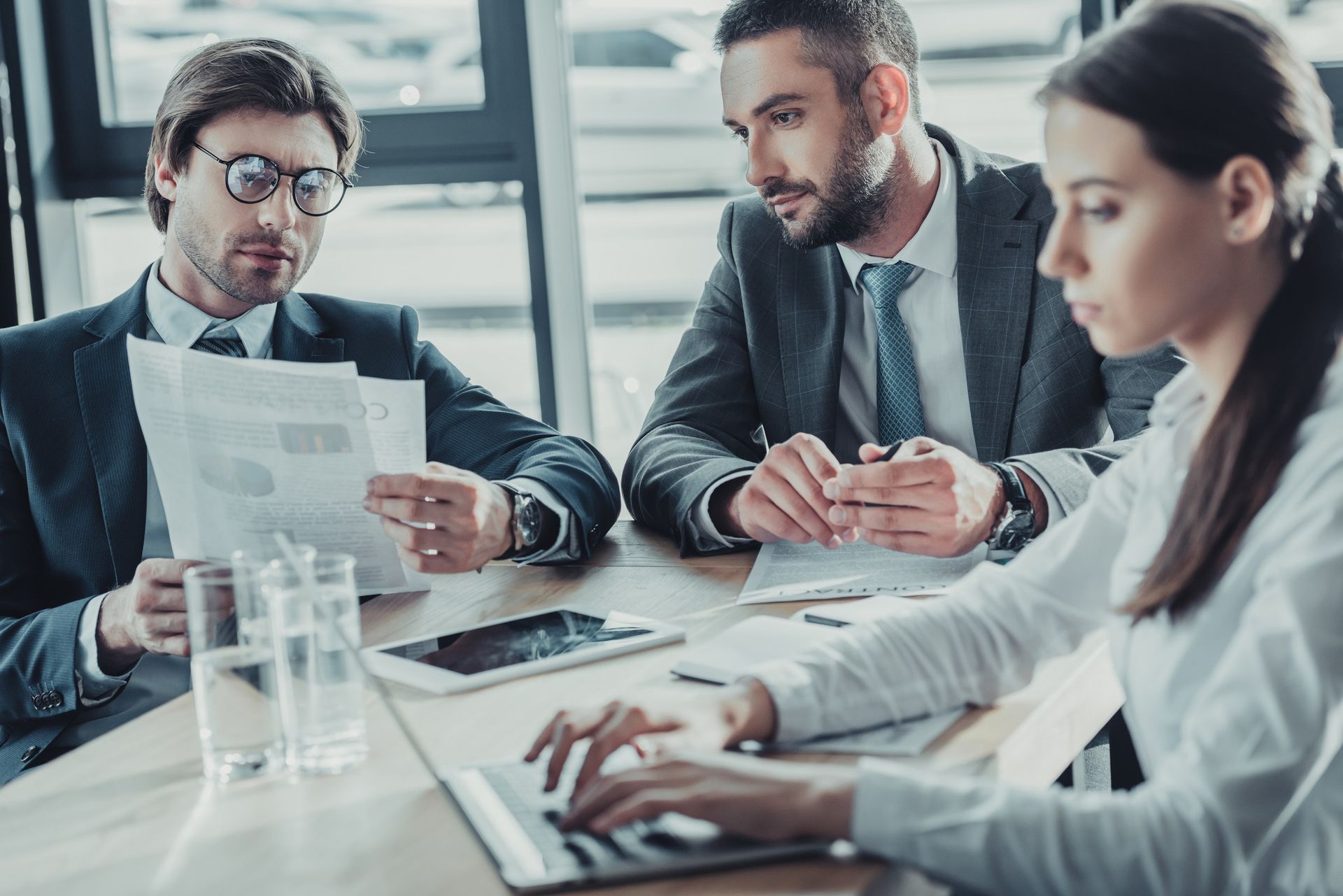 Three businesspeople review documents and use a laptop at a desk.