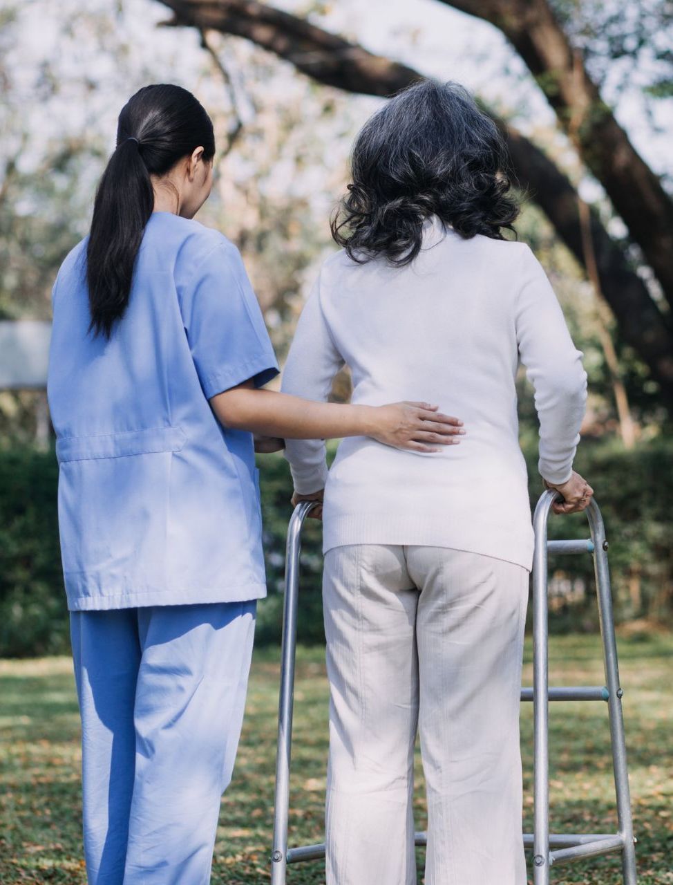 Nurse in blue guides elderly woman using a walker outdoors, touching her back for support.
