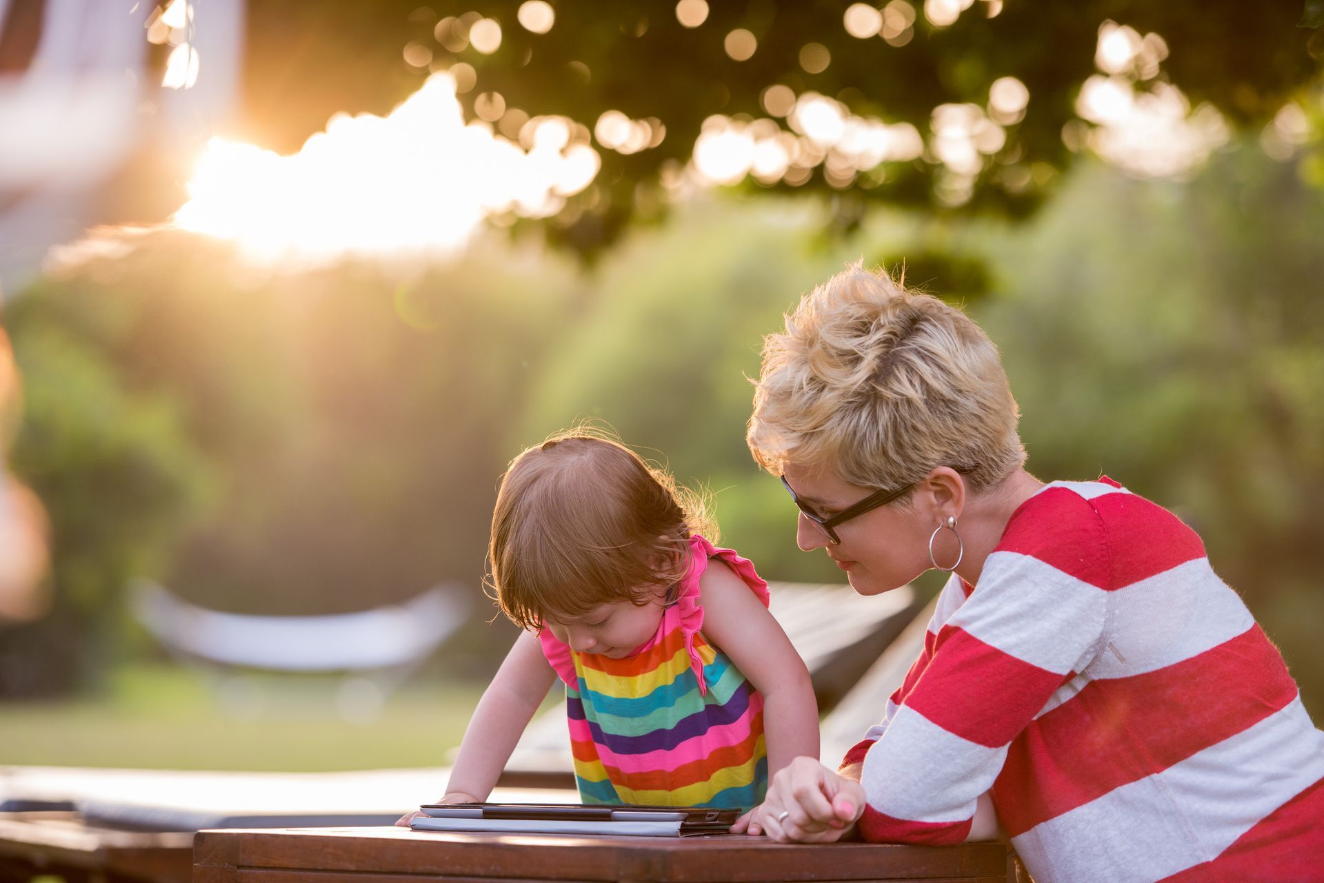 A woman with short blonde hair and a striped shirt looks at a toddler in a colourful dress at a table outdoors.