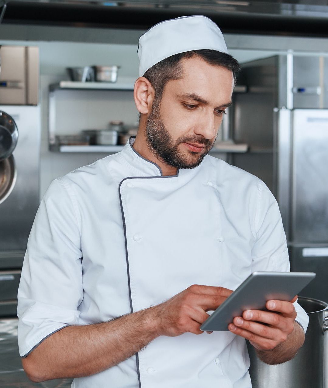 Chef in white uniform and hat using a tablet in a commercial kitchen.