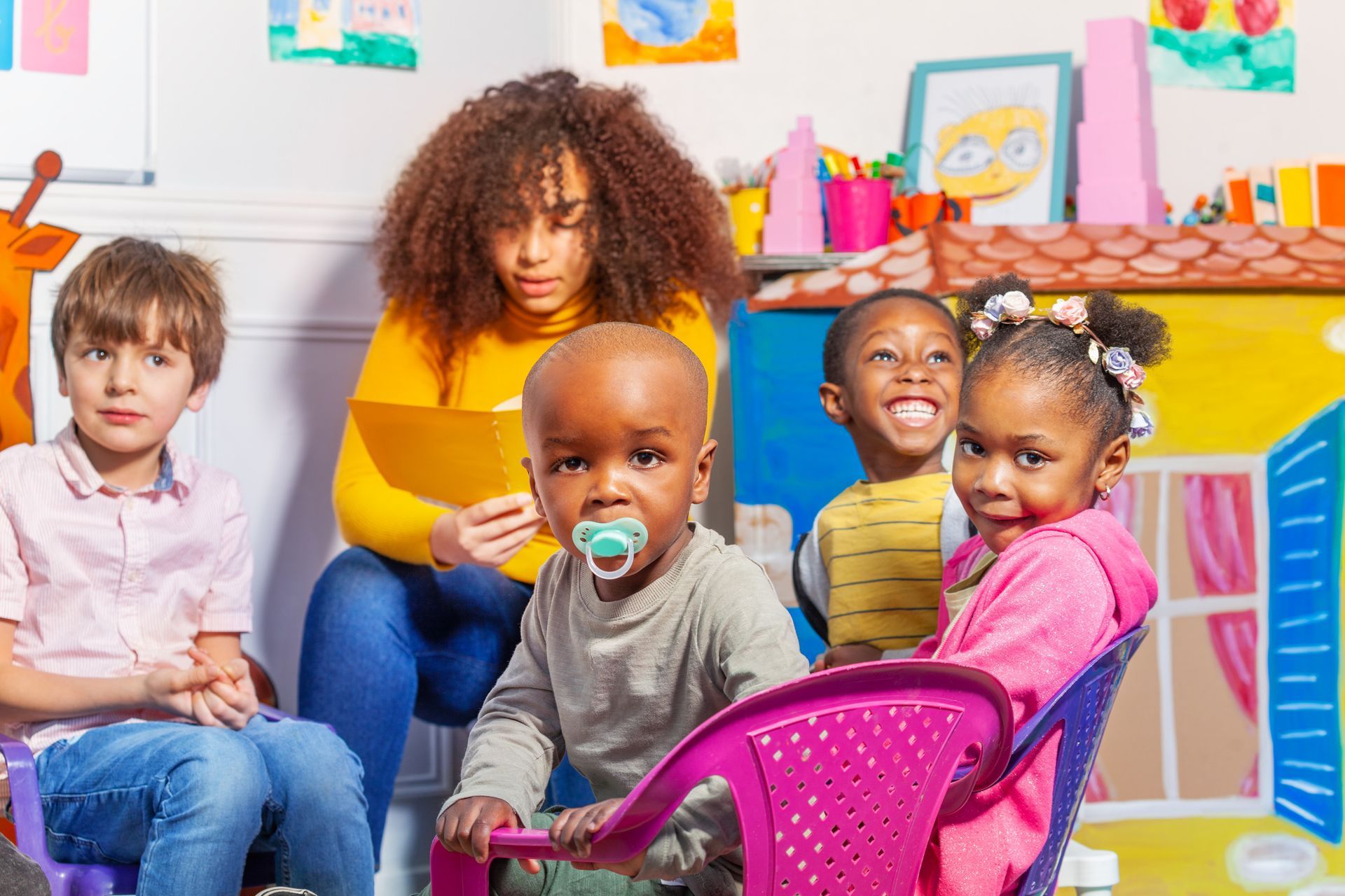 Children and teacher in a colorful playroom; the teacher reads to the group; kids smile.