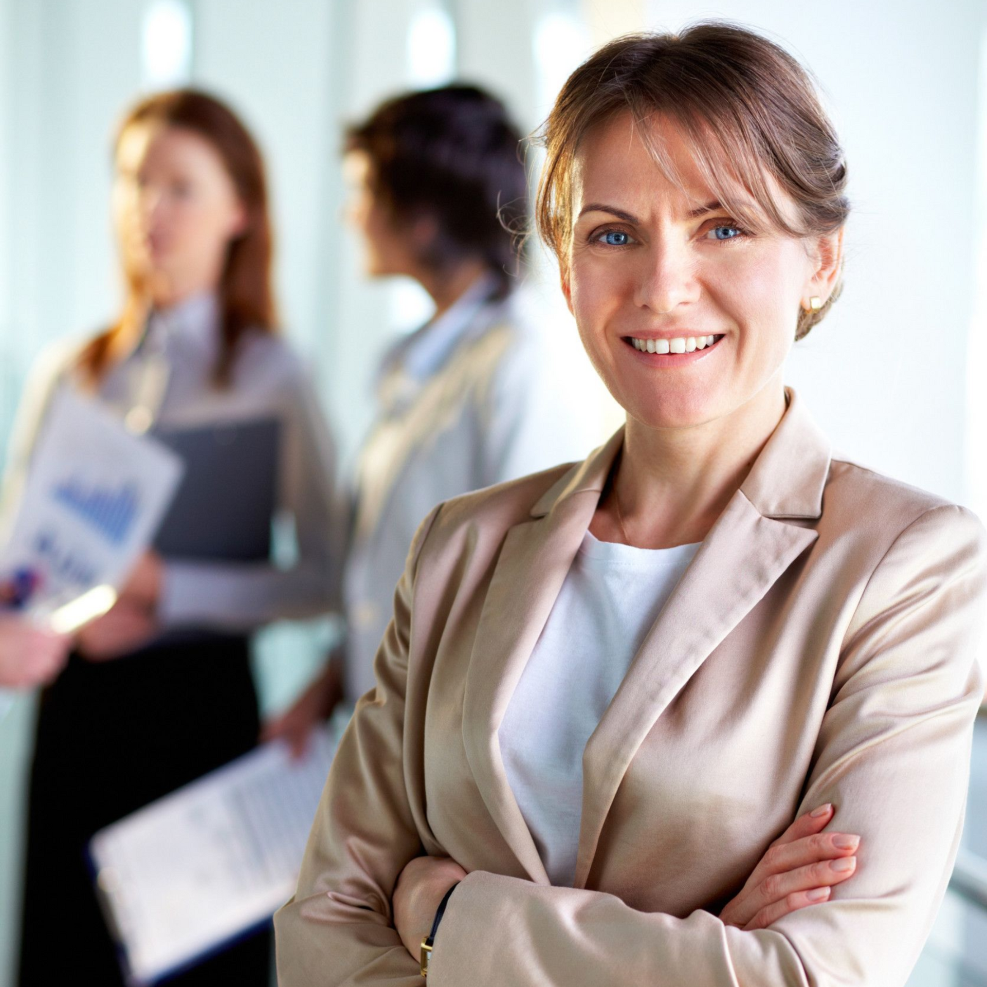 Woman in tan blazer smiles, arms crossed, in office setting with coworkers.