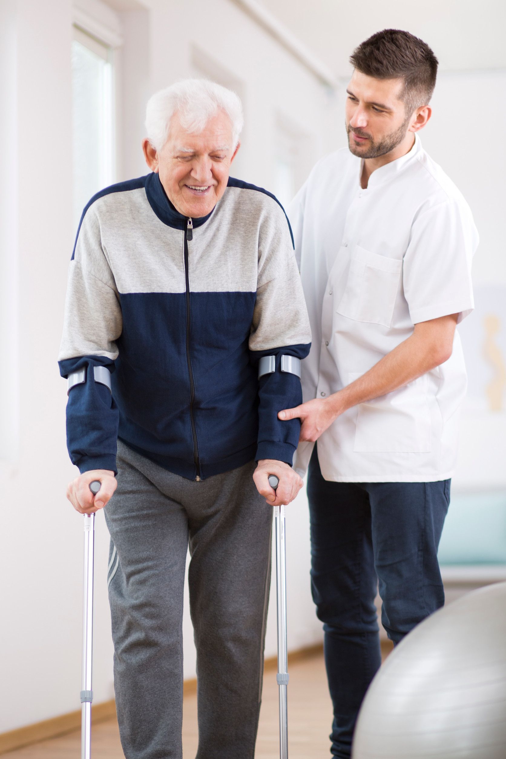 Elderly man using crutches, assisted by a healthcare professional, indoors.
