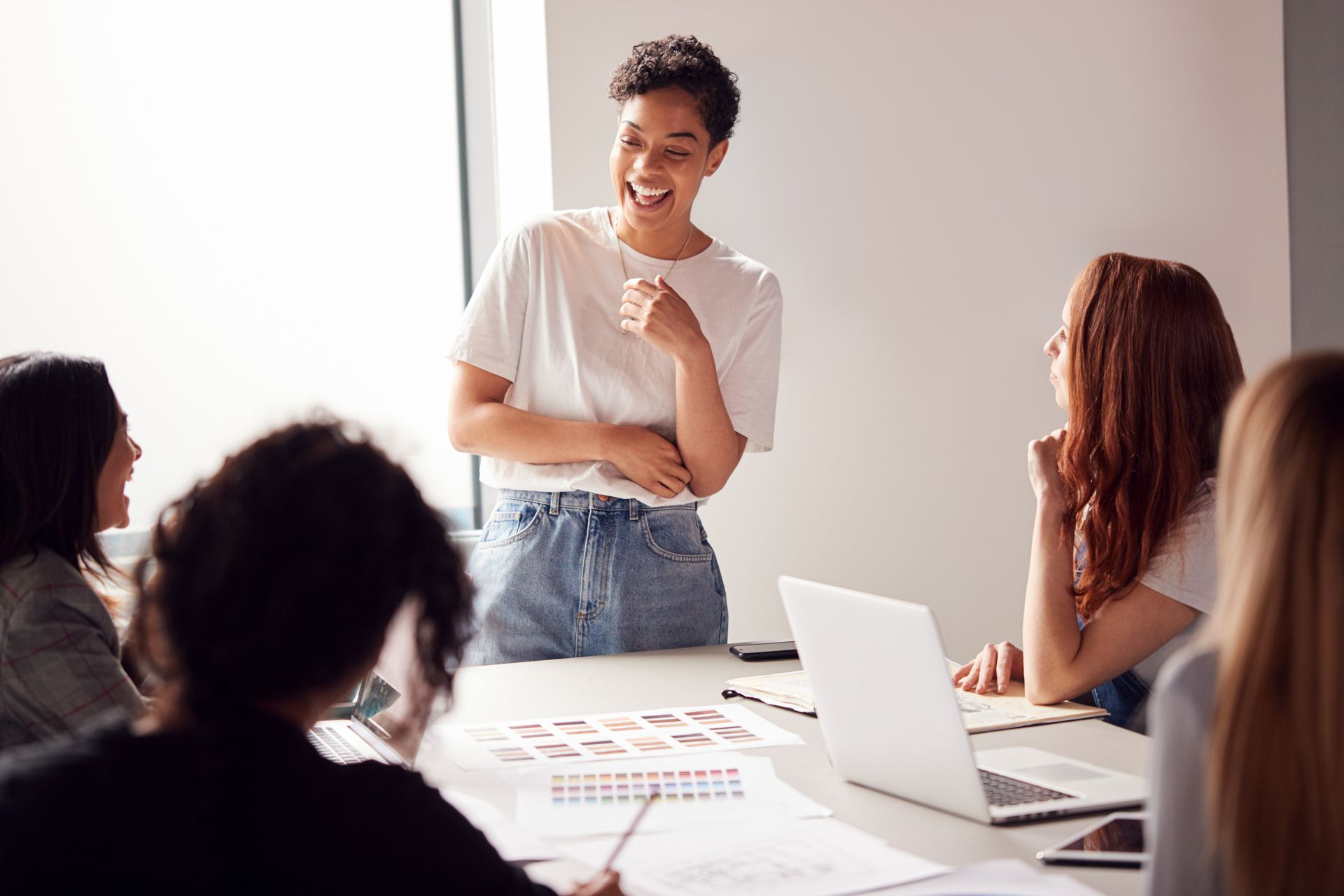 Woman in a white shirt speaking to colleagues at a table with laptops and papers.