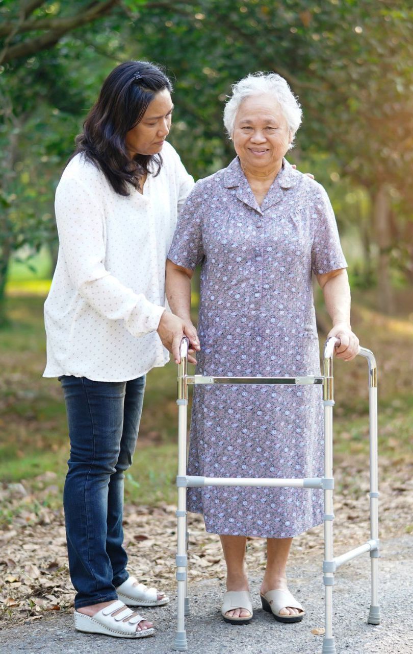 Woman assisting elderly woman using a walker outdoors; both smiling.
