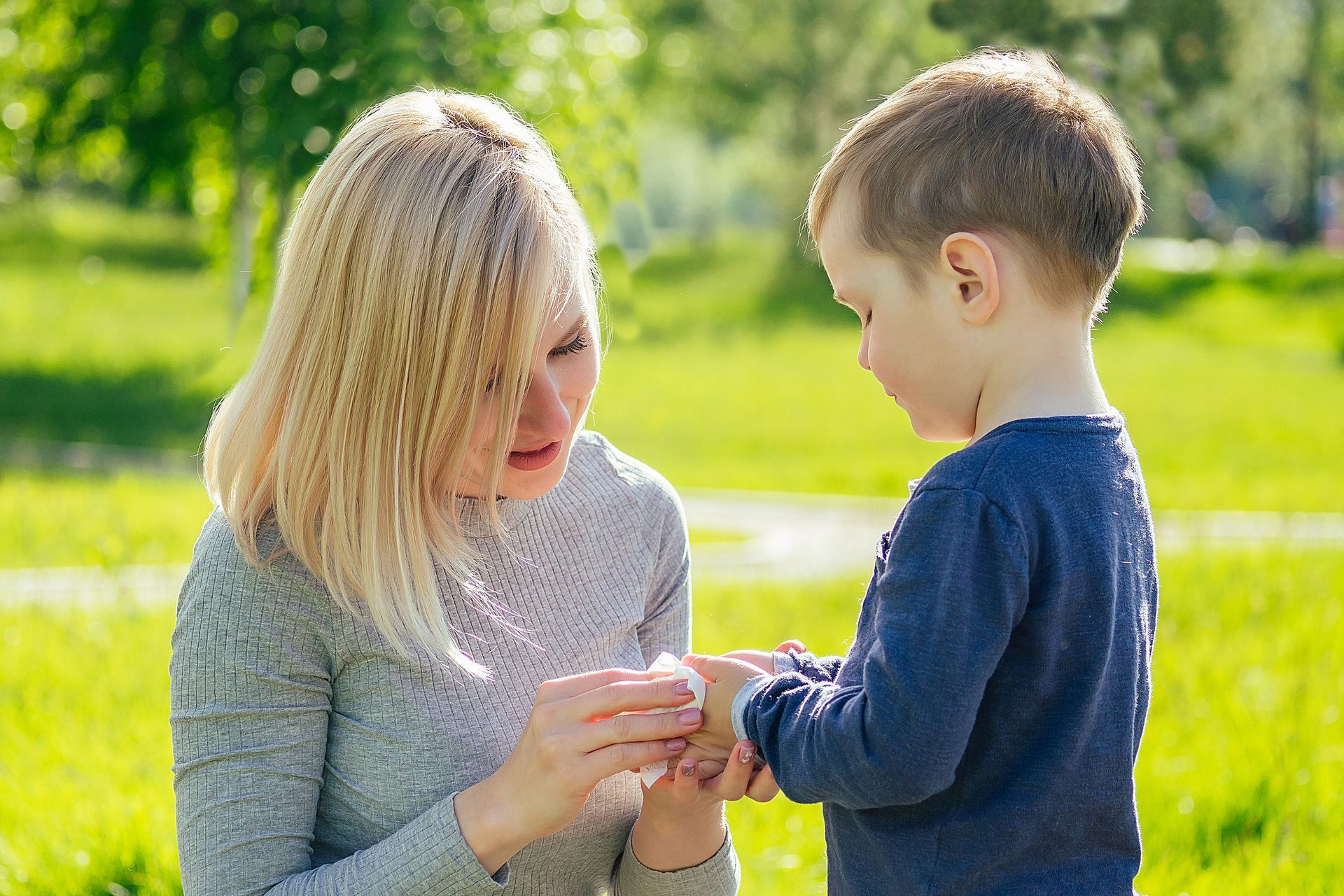 Woman putting a bandage on a boy's wrist in a sunny park setting.